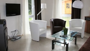 Un salon moderne avec deux fauteuils blancs, une table basse en verre, un fauteuil en cuir noir, un téléviseur à écran plat et une grande fenêtre avec des rideaux.