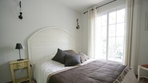 Un dormitorio con cama, mesita de noche, lámpara, ventana con cortinas y un aplique de pared.