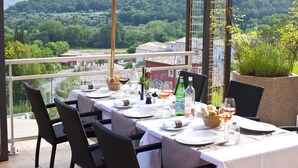 Mesa de comedor servida con platos, vasos y botellas, con vistas a un paisaje de un pueblo y un bosque.