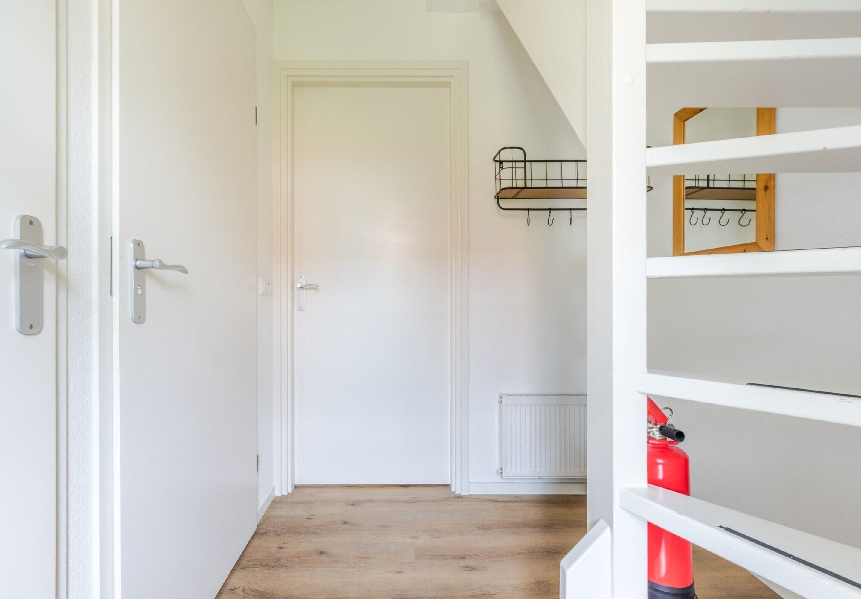 Un couloir avec une porte blanche, un radiateur et une étagère sur laquelle se trouve un objet rouge.