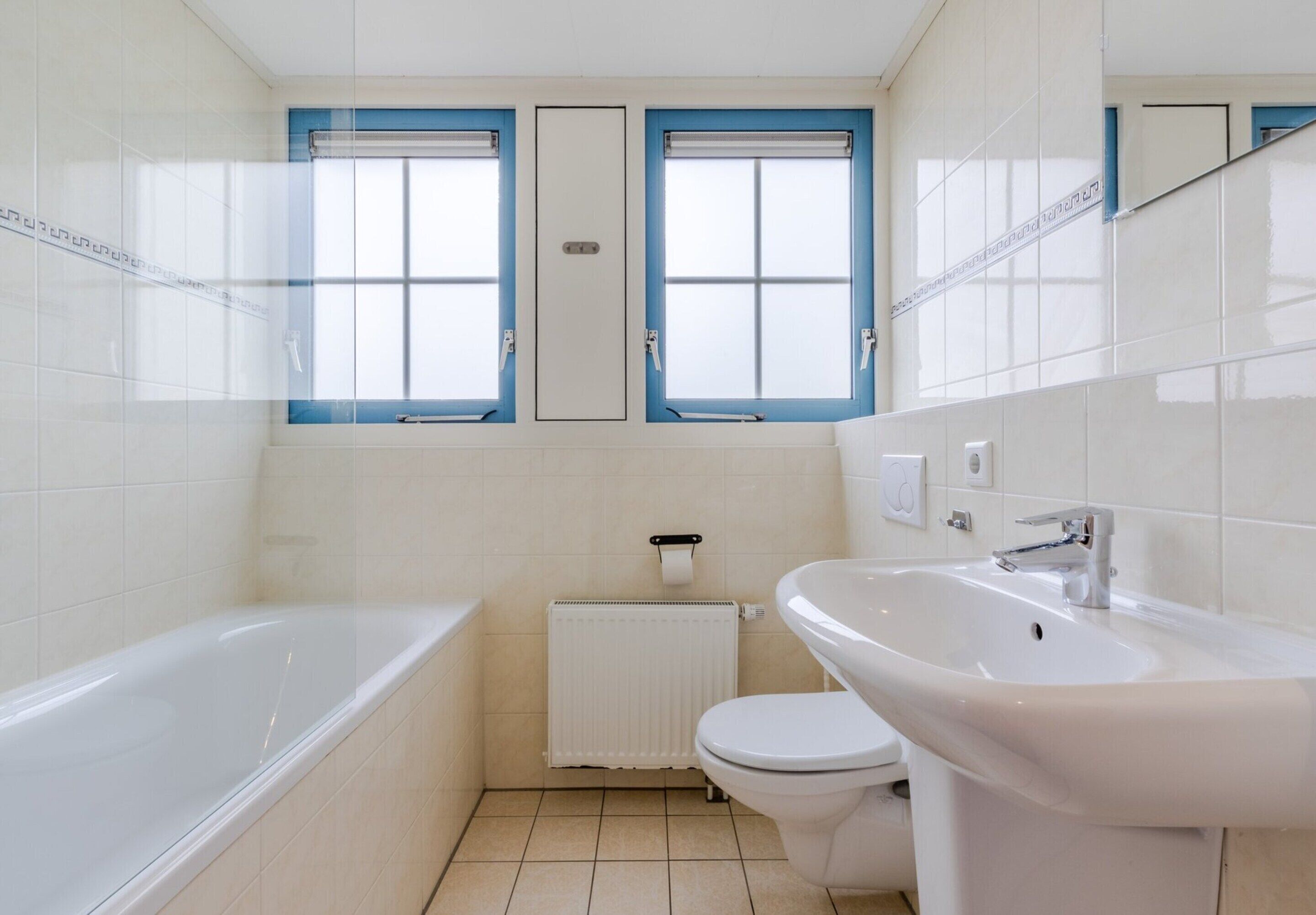 Une salle de bain avec une baignoire, des toilettes, un lavabo et deux fenêtres aux cadres bleus.