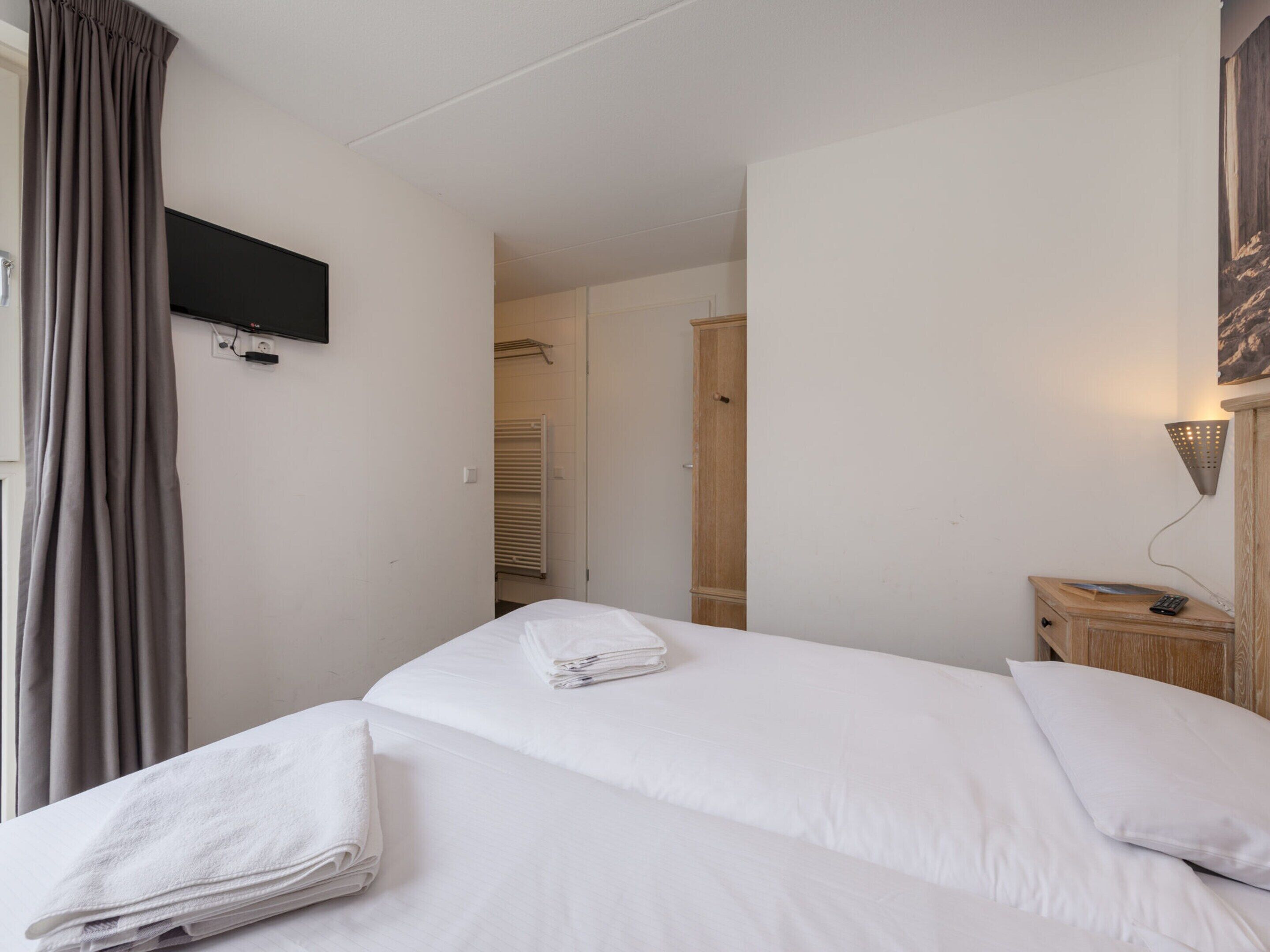 A hotel room with a bed, a TV mounted on the wall, a wooden nightstand, and a door leading to a bathroom.