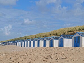 Una serie di casette sulla spiaggia, con porte blu e pareti bianche, allineate lungo una spiaggia sabbiosa con dune sullo sfondo.