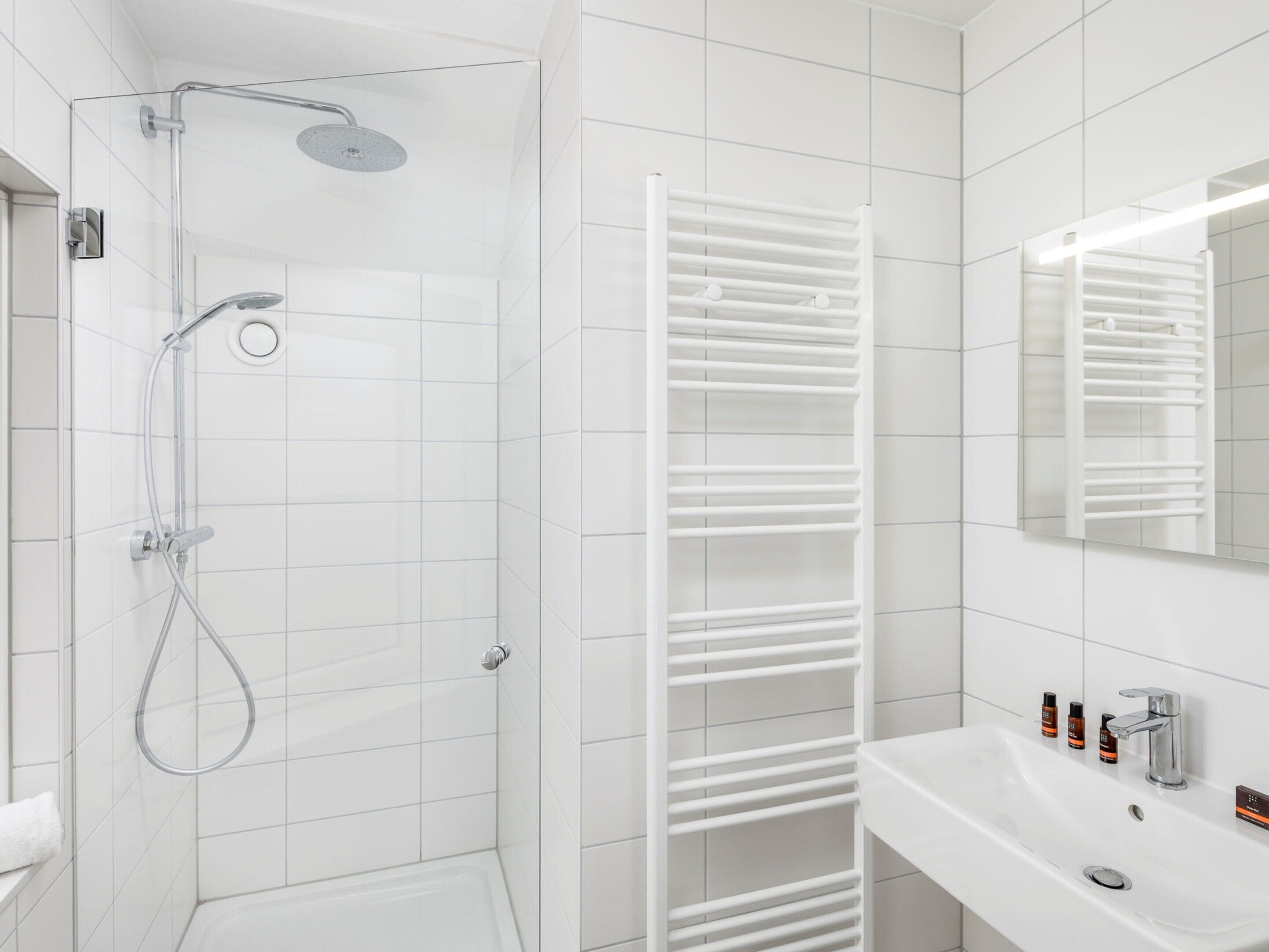 Ein modernes Badezimmer mit einer gläsernen Duschkabine, einer weiß gefliesten Wand, einem weißen Handtuchhalter und einem Waschbecken mit Spiegel.