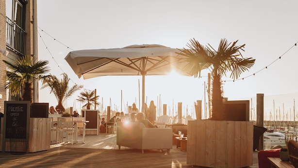 An outdoor seating area with wooden tables and chairs, a large umbrella, and a view of the sea with boats in the background. - Chalet Near the Beach With Dishwasher (Kamperland)