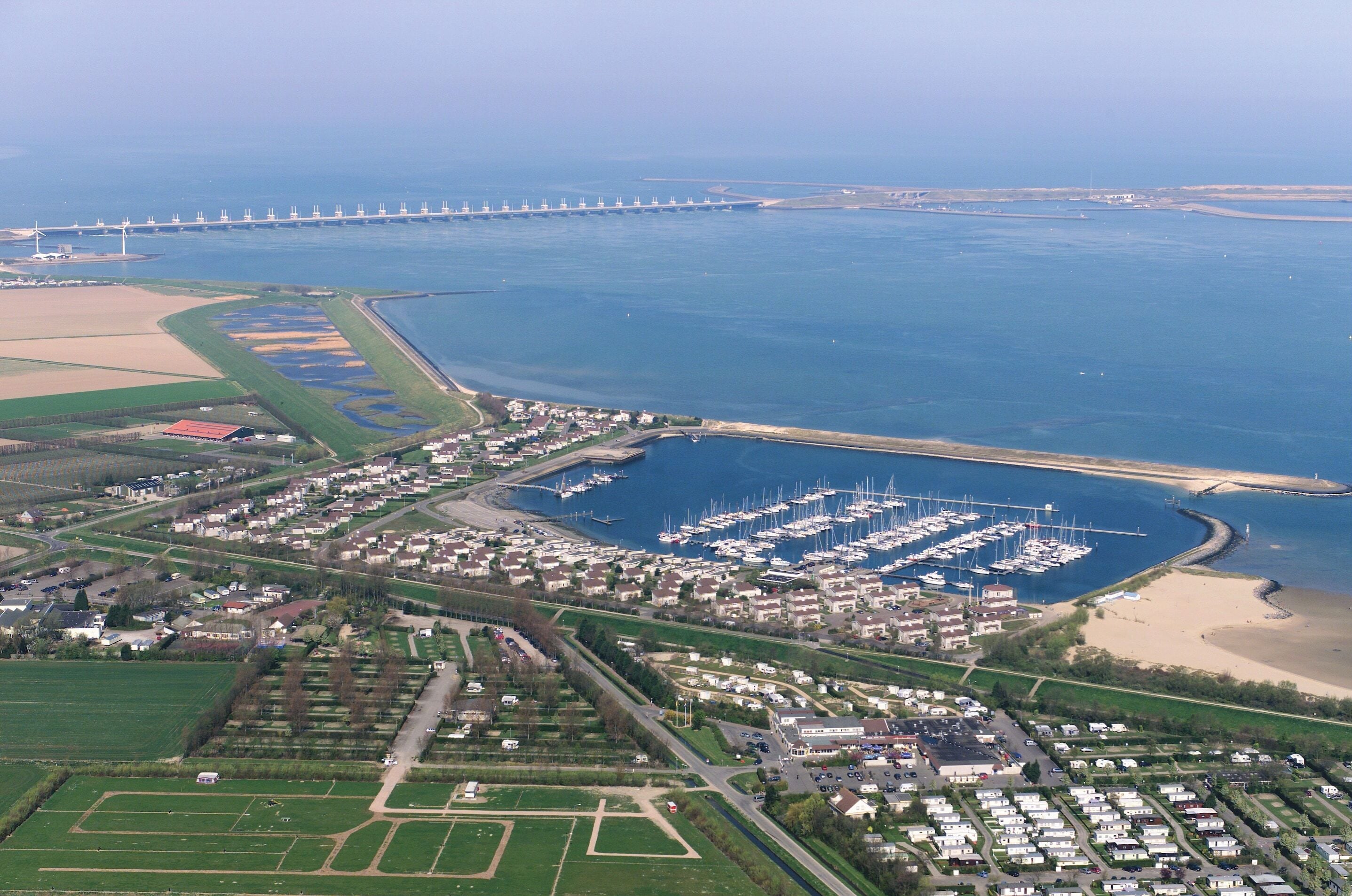 An aerial view of a coastal marina with boats, a bridge in the background, and residential areas with houses and roads.