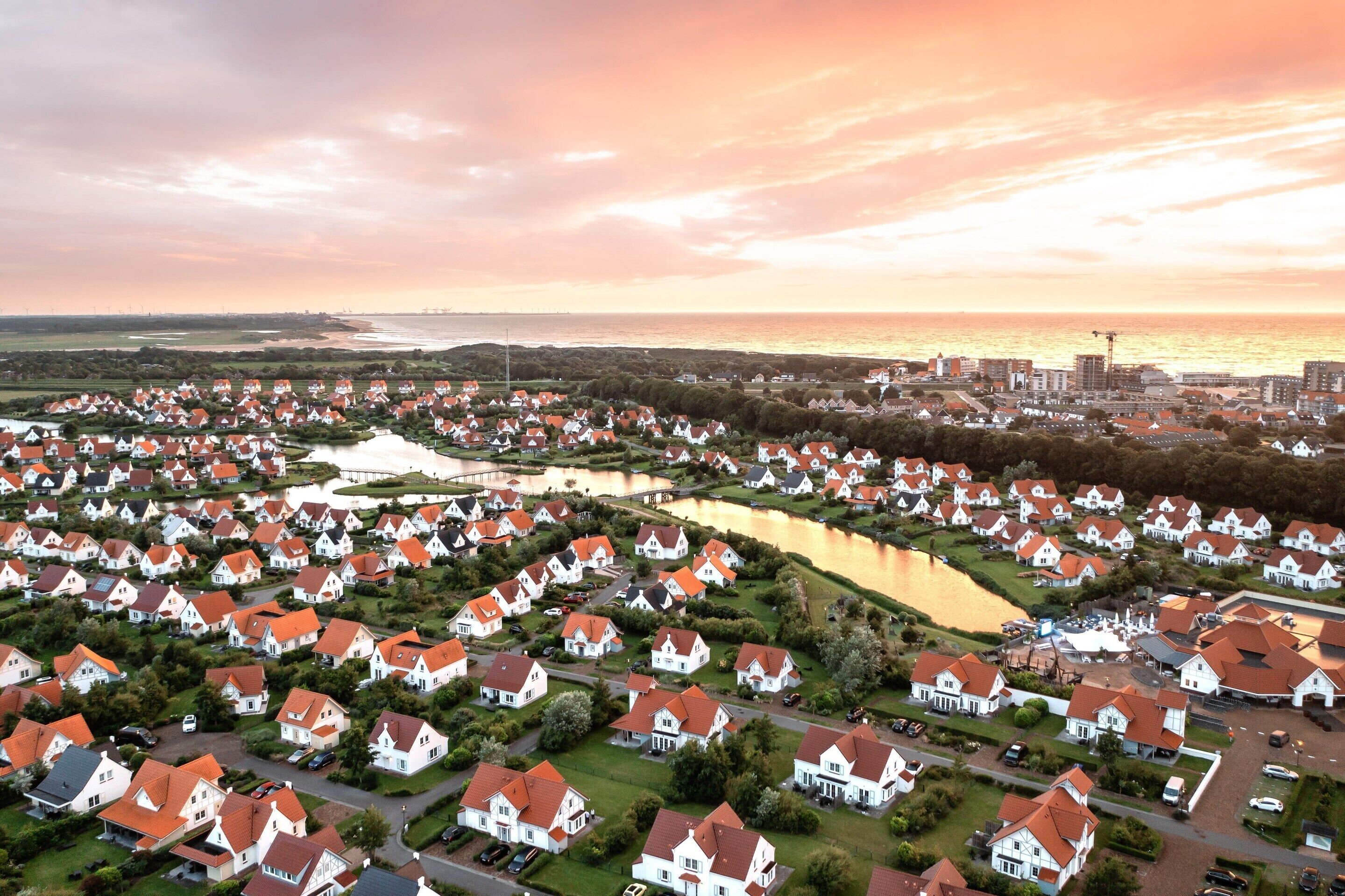 Vue aérienne d’un quartier résidentiel avec des maisons, une étendue d’eau et une silhouette de ville en arrière-plan, au coucher du soleil.