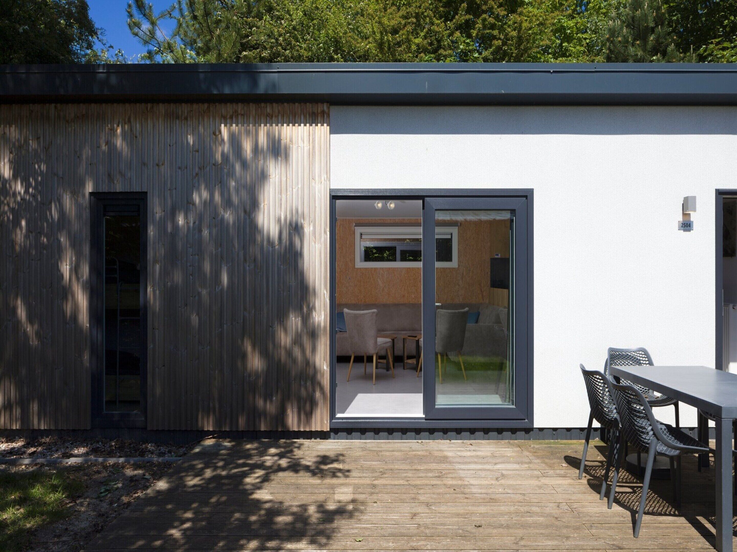 Ein modernes Haus mit einer Holzterrasse, einer Schiebetür zu einem Wohnbereich und einem kleinen Tisch mit Stühlen im Freien.