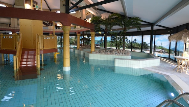 Una piscina interior con suelo de baldosas, un tobogán de madera y una zona con techo de paja.