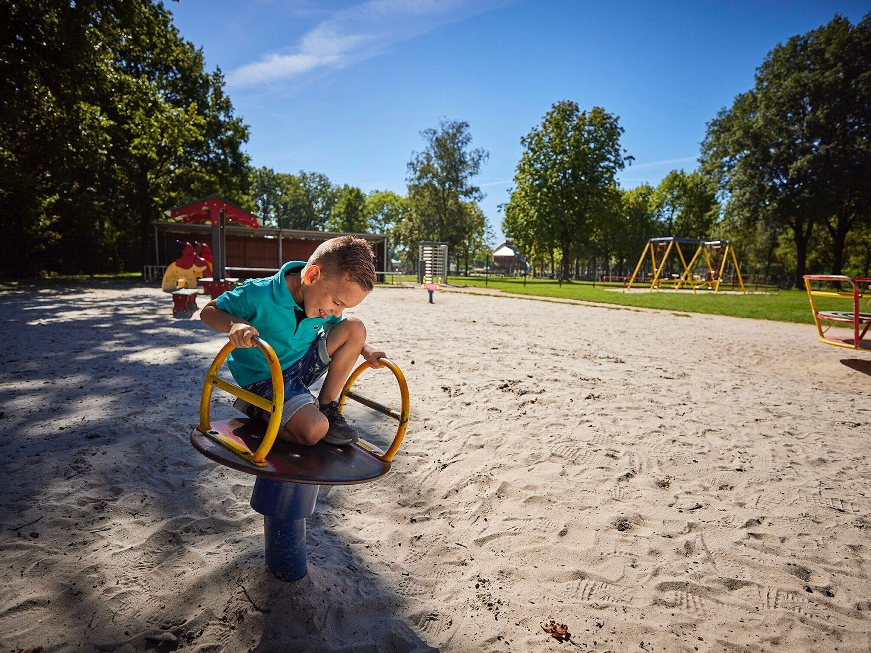 Ein Kind schaukelt auf einer Wippe im Park. Im Hintergrund sind Sand, Bäume und Spielgeräte zu sehen.