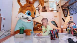 Un bambino disegna a un tavolo con un grande mascotte a forma di coniglio e diversi articoli da disegno.