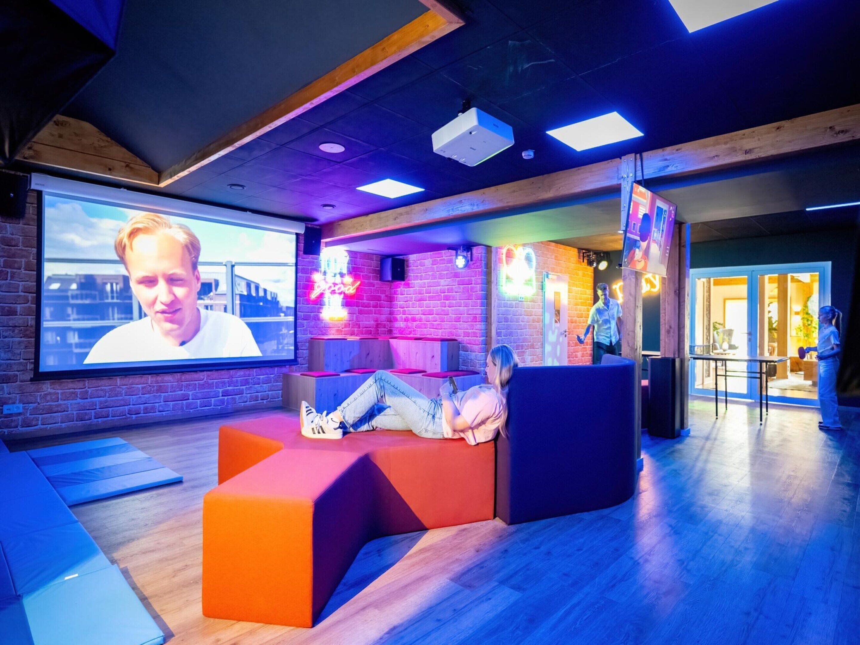A modern lounge area with a large screen, comfortable seating, and a brick wall backdrop.