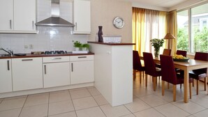 Une cuisine moderne avec des armoires blanches, une cuisinière à gaz et une hotte en acier inoxydable. On y trouve un coin repas avec une table en bois et des chaises rouges. Une horloge est visible au mur.