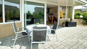 Une terrasse avec une table ronde et des chaises, une porte vitrée donnant sur un coin repas et un mur en briques.