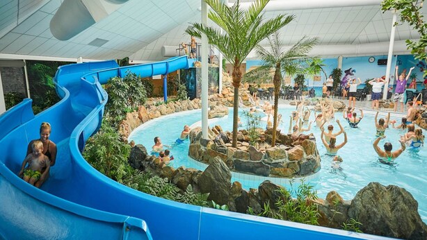 Un parco acquatico al coperto con uno scivolo d'acqua blu, una piscina centrale circondata da rocce e palme, e persone che nuotano e si divertono.