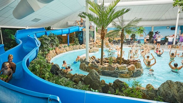 Un parc aquatique intérieur doté d’un toboggan bleu, de plusieurs piscines et de palmiers.