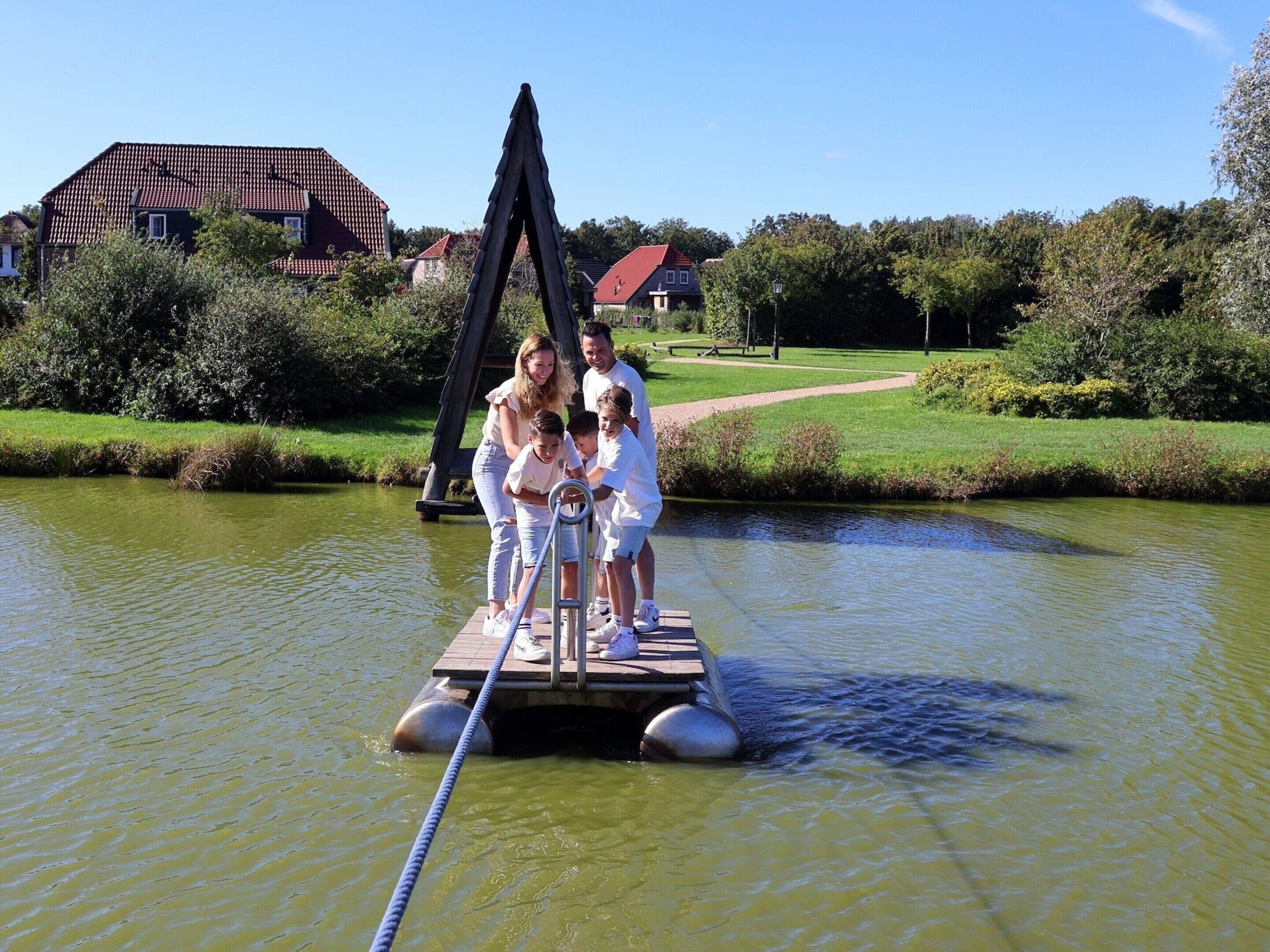A family on a floating platform in a calm body of water, surrounded by greenery and houses in the background.