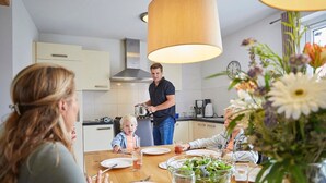 Une cuisine moderne avec une table à manger dressée pour le repas, une personne qui cuisine et un vase rempli de fleurs sur la table.