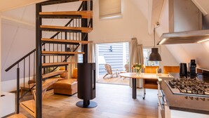 Una cocina moderna con estufa a gas, un comedor con mesa redonda y sillas, una escalera con escalones de madera y barandillas negras, y un ventanal con cortinas.