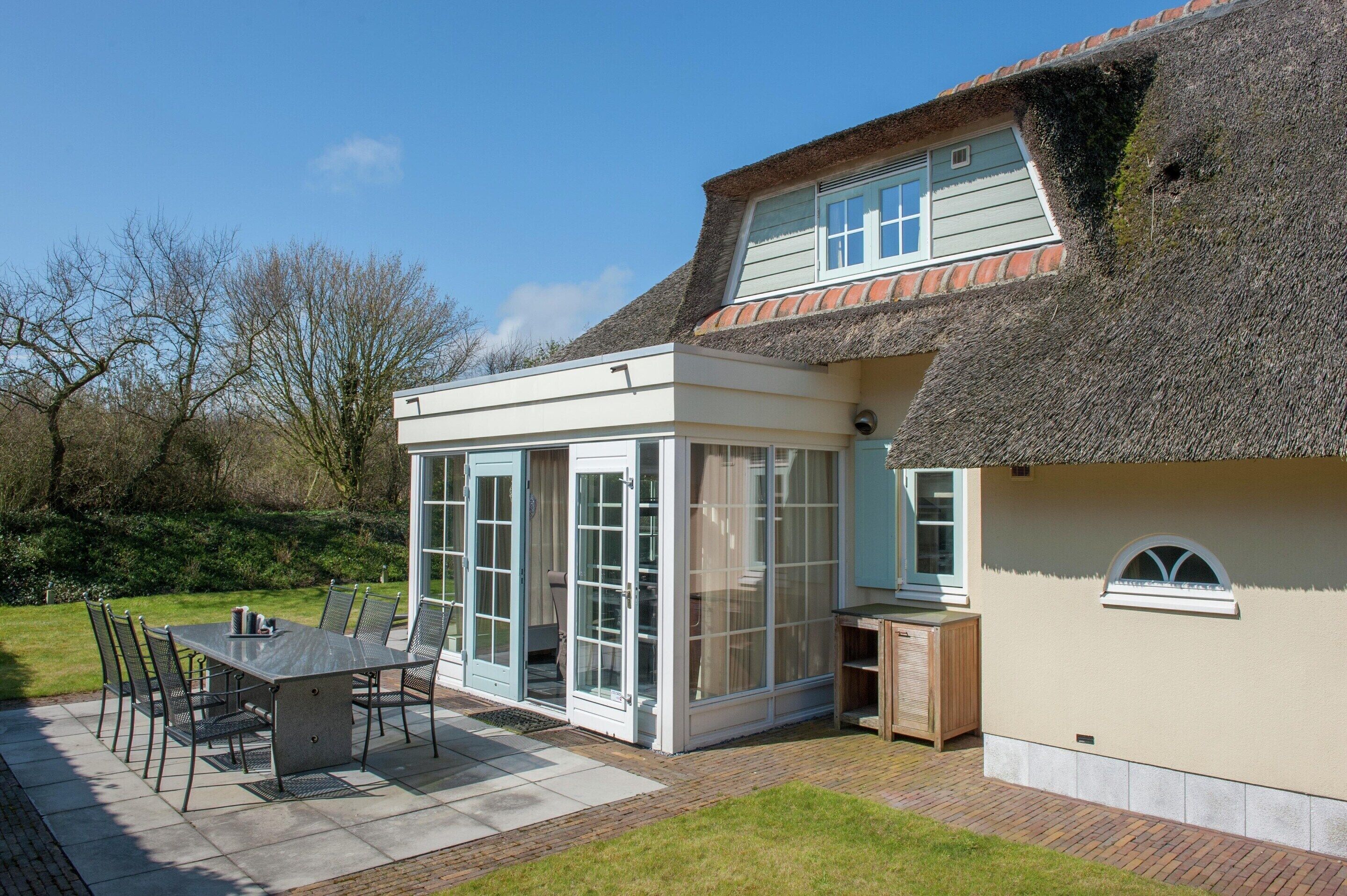 A house with a thatched roof, a conservatory, and a patio with a table and chairs.
