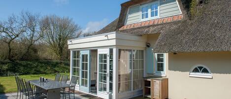 A house with a thatched roof, a conservatory, and a patio with a table and chairs.