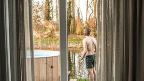 Iemand staat bij een houten terras met uitzicht op een vijver, waar een hottub en een houten bank zichtbaar zijn.