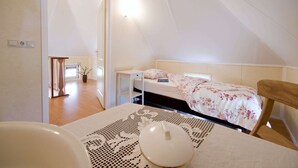 Quarto com cama, mesa de cabeceira e uma cadeira. Há uma janela com cortinas, uma porta que dá para outro cômodo e uma pequena mesa com um vaso.