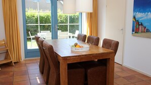 Ein Esszimmer mit einem Holztisch, sechs bequemen braunen Stühlen, einer weißen Schüssel mit Früchten und einem großen Fenster mit Vorhängen.