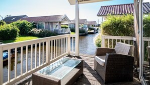 A covered balcony with wicker furniture, a glass table, and a view of a canal with houses in the background. - Cozy Chalet Right on the Water in Friesland (Akkrum)