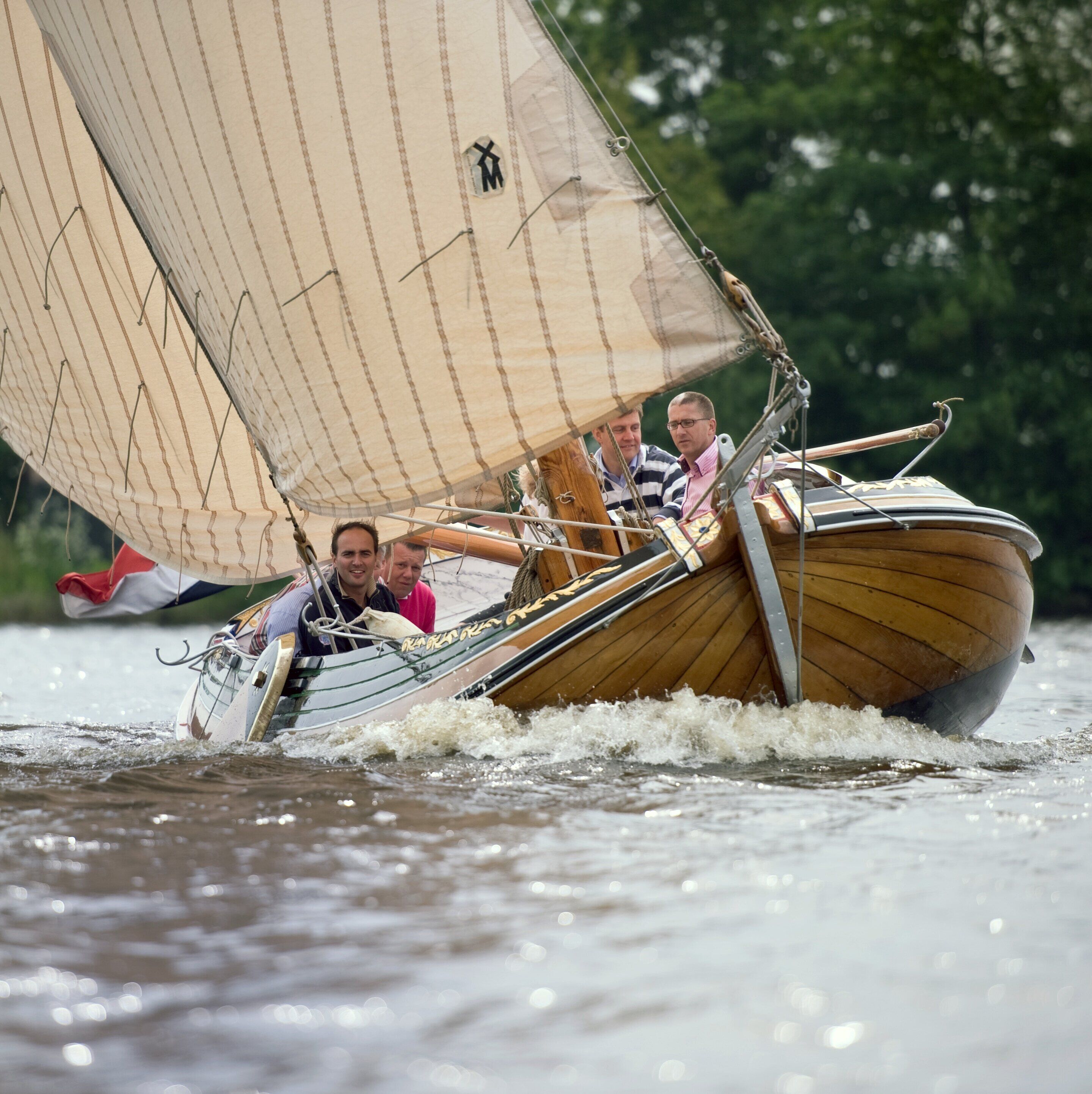 Een houten boot met zeil, bemand door drie personen, op het water.