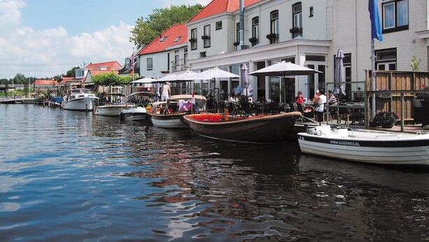 A waterfront area with boats docked along the shore, buildings with red roofs, and outdoor seating areas with umbrellas. - Cozy Chalet Right on the Water in Friesland (Akkrum)