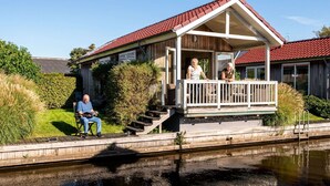 A wooden house with a red roof, a porch, and a small deck by a canal. - Cozy Chalet Right on the Water in Friesland (Akkrum)