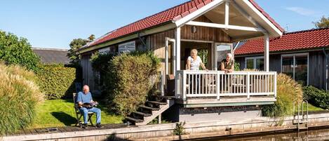 A wooden house with a red roof, a porch, and a small deck by a canal.