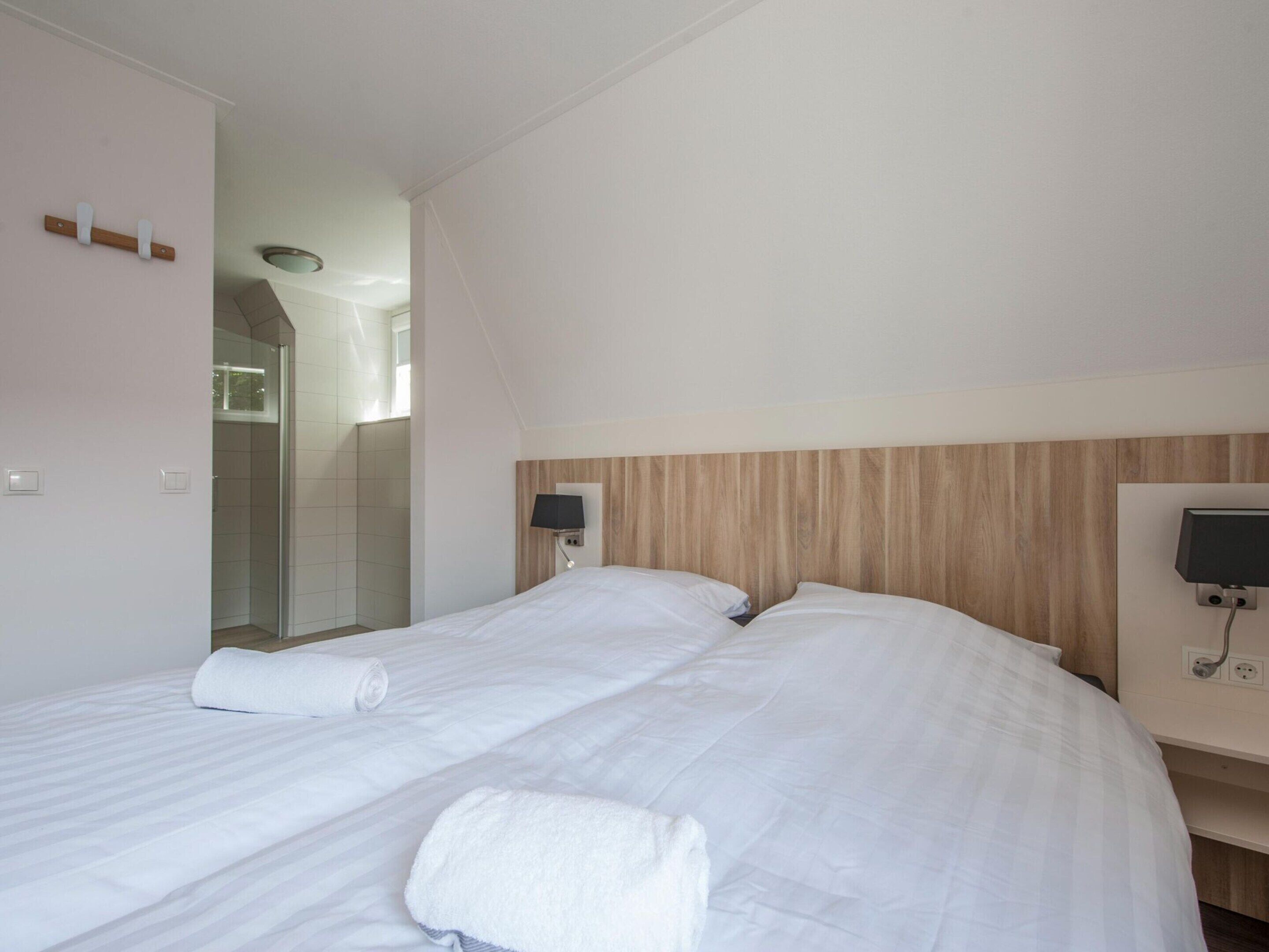 Habitación de hotel con dos camas, un cabecero de madera, una mesita de noche con lámpara y un área de ducha visible a través de una puerta de vidrio.