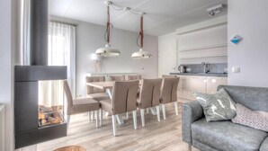 Ein modernes Wohnzimmer mit grauem Sofa, einem Essbereich mit Holztisch und -stühlen, einer Küche mit weißen Schränken und einem Kamin mit Holzscheiten.