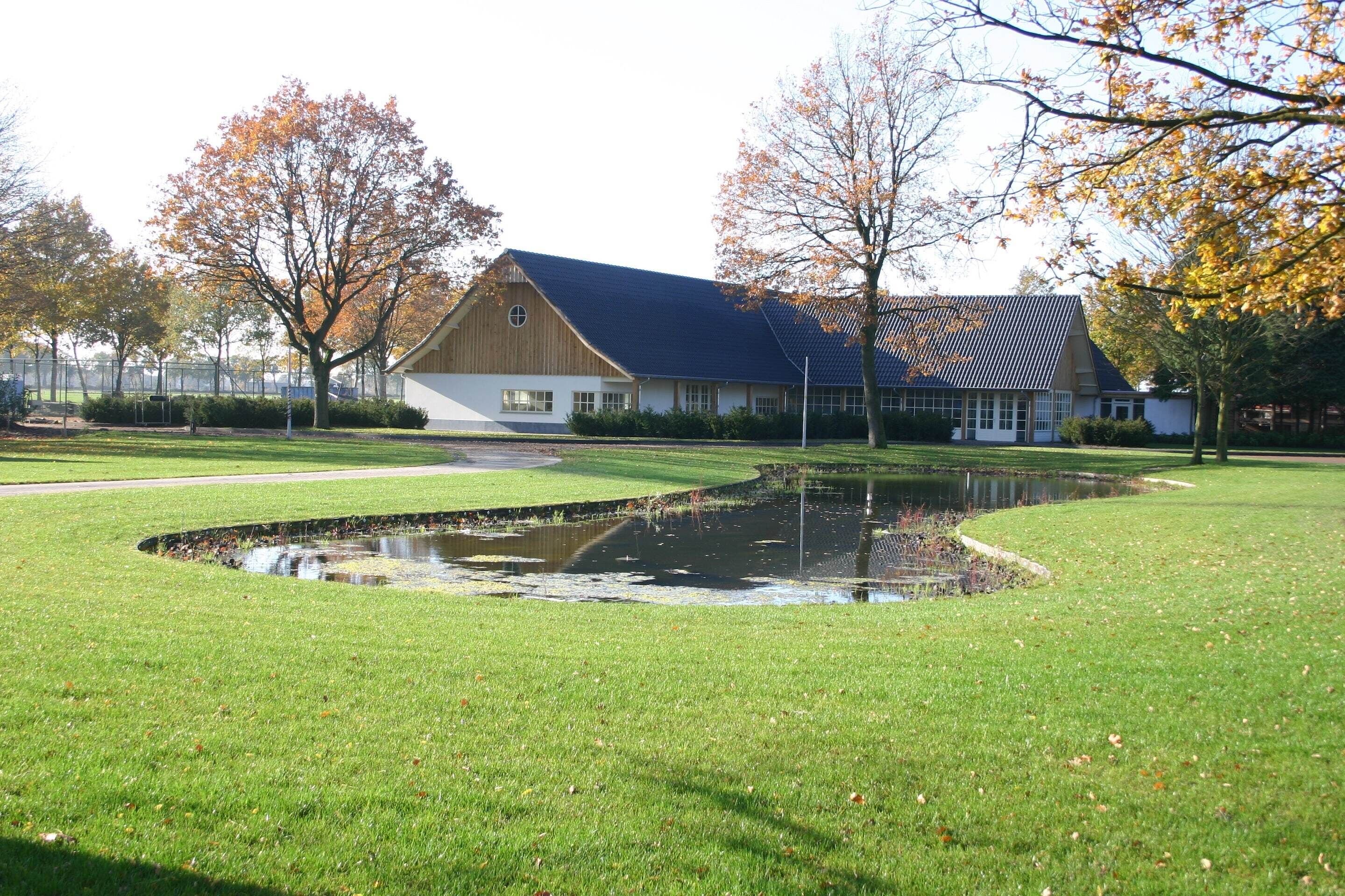 Un bâtiment situé devant un grand étang, entouré de pelouses bien entretenues et d’arbres aux feuilles d’automne.