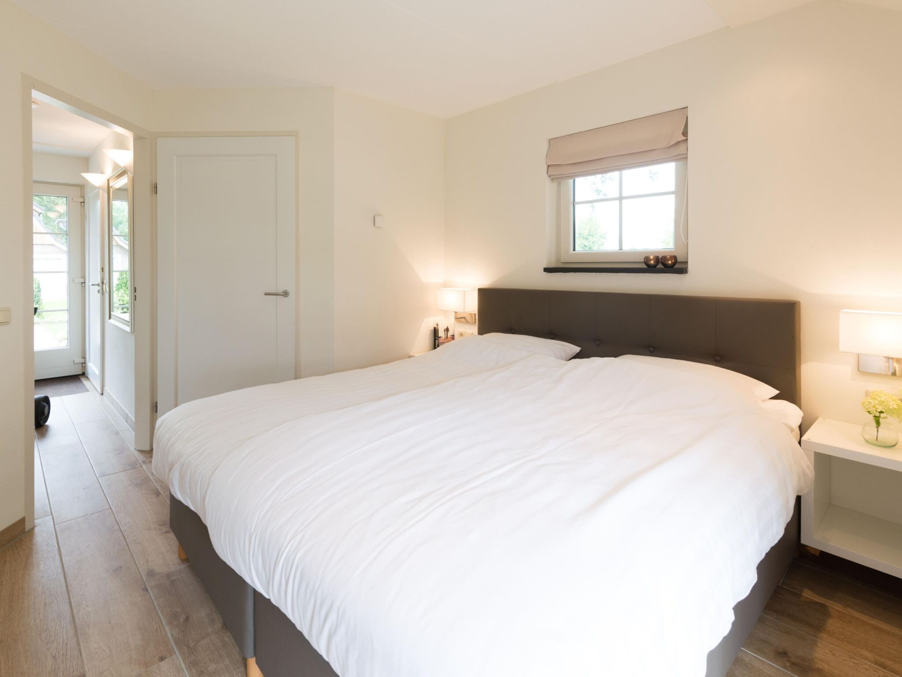 A bedroom with a large bed, a window with a view, a door, and a bedside table with a lamp.