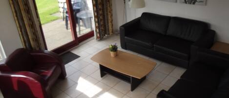 A living room with a black sofa, a red armchair, and a wooden coffee table. There is a sliding glass door leading to a patio with outdoor furniture.