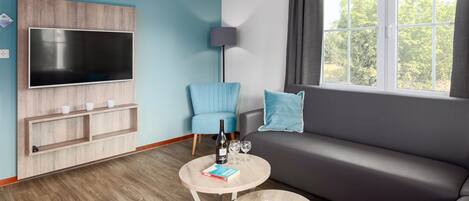 A modern living room with a grey sofa, a blue armchair, a TV mounted on a wooden panel, a small round table with a book and glasses, and a window with curtains.