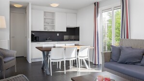 A modern kitchen with white cabinets, a dining table with chairs, and a sofa in a bright living area with large windows. - Bungalow in Zeebad Near the Beach (Breskens)