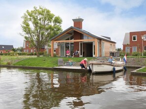 A modern house with a glass door, a small boat docked at the water's edge, and a grassy area with a tree.