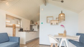 A modern, well-lit living area with a sofa, coffee table, and a dining table with chairs. A kitchen is visible in the background. - Ameland Lodge w/ Veranda (Hollum)