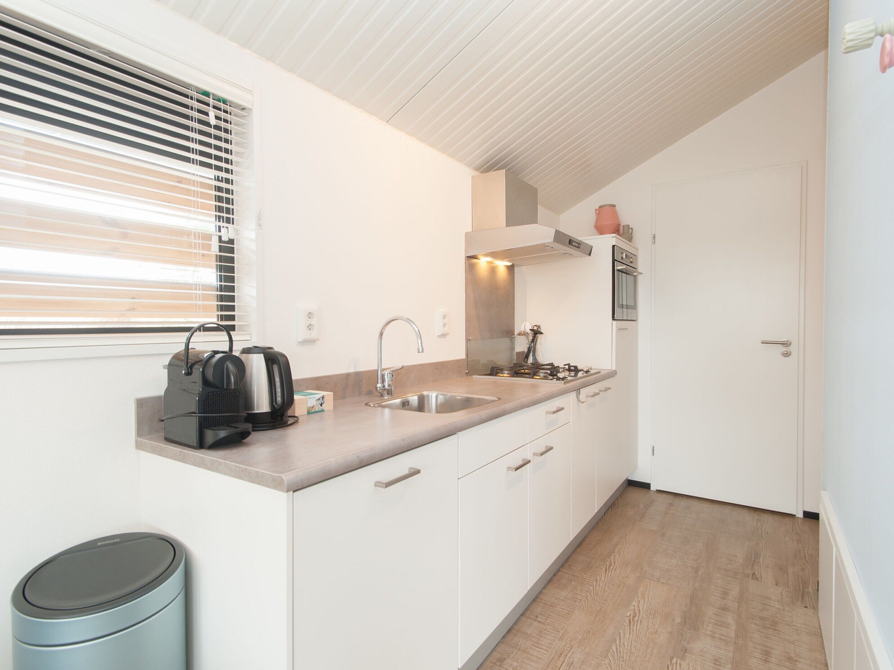 Una cocina compacta con lavabo, estufa y hervidor, con armarios blancos y suelo de madera.