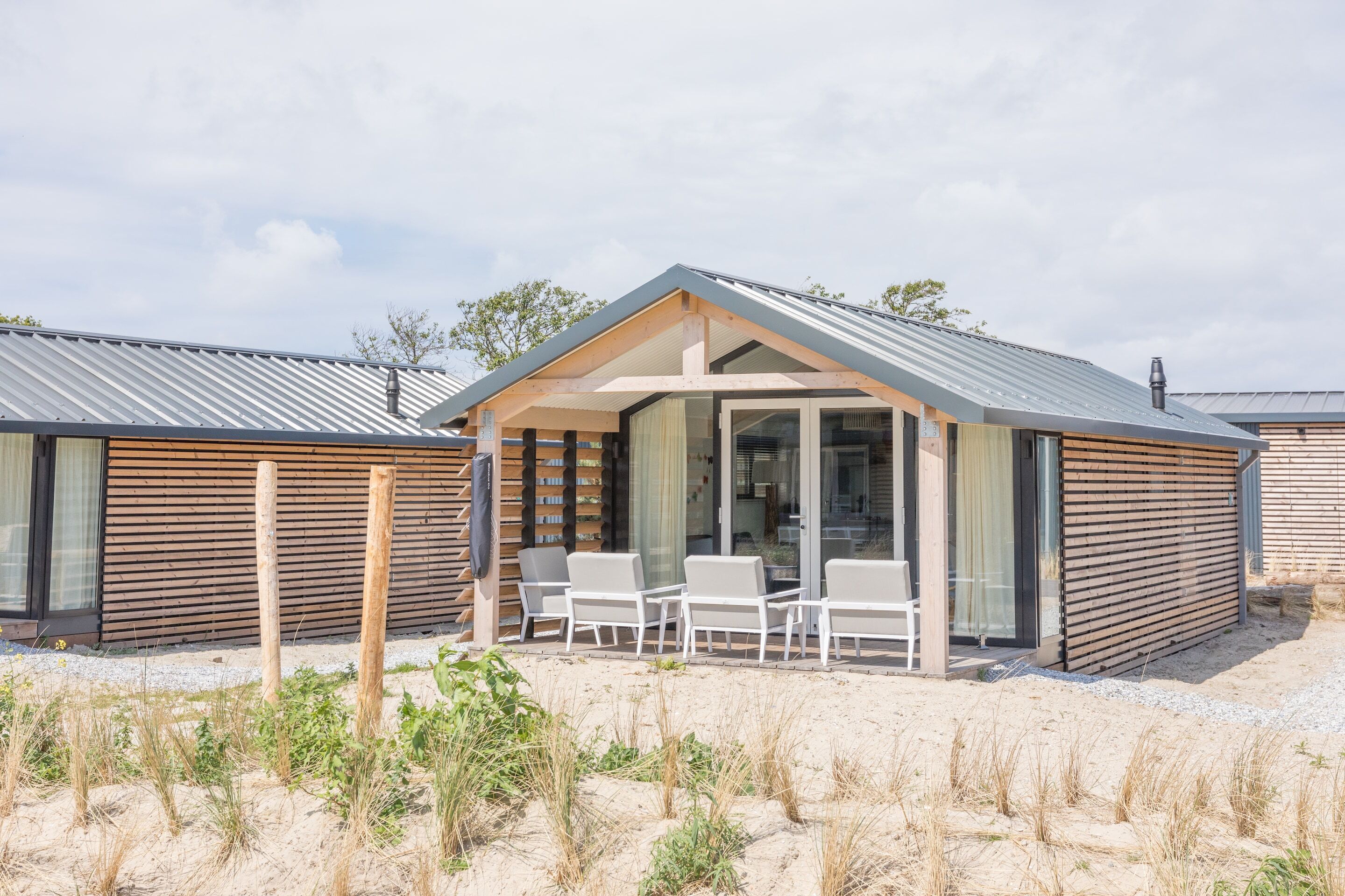 Ein modernes Strandhaus mit überdachter Terrasse, Sitzgelegenheiten im Freien und einer sandigen Landschaft.