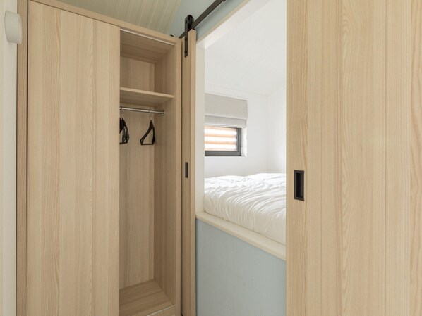 A wooden wardrobe with a sliding door, a hanging rack, and a small window above a bed. - Ameland Lodge w/ Veranda (Hollum)