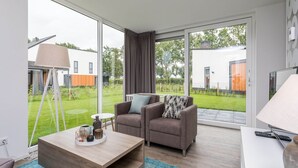 Uma sala de estar moderna com sofá, mesa de centro e luminária. Há uma janela grande que oferece vista para um jardim e edifícios vizinhos.