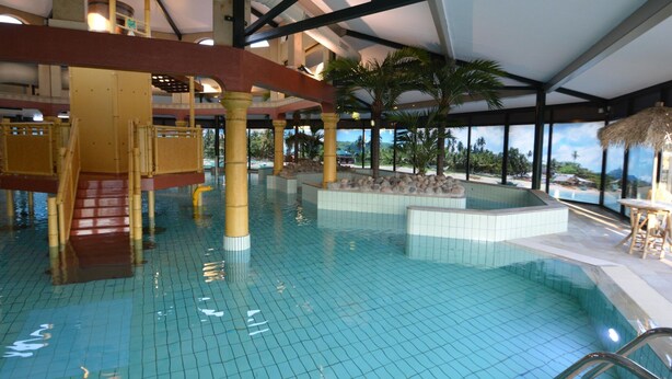 Una piscina interior con suelo de baldosas, un tobogán de madera y una zona con techo de paja.