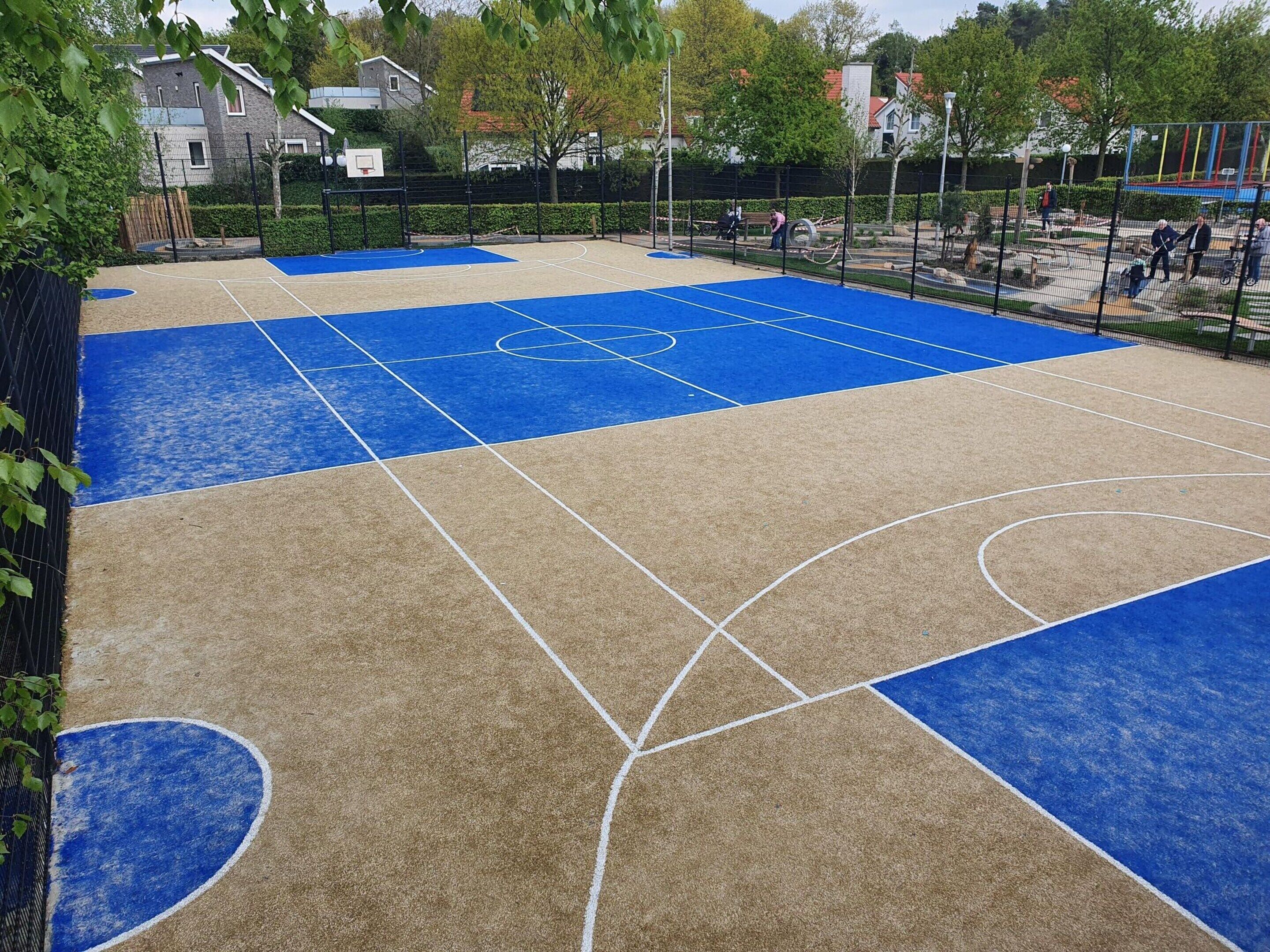 Ein Basketballplatz im Freien mit blau-beiger Spielfläche, umgeben von einem Zaun und Bäumen.