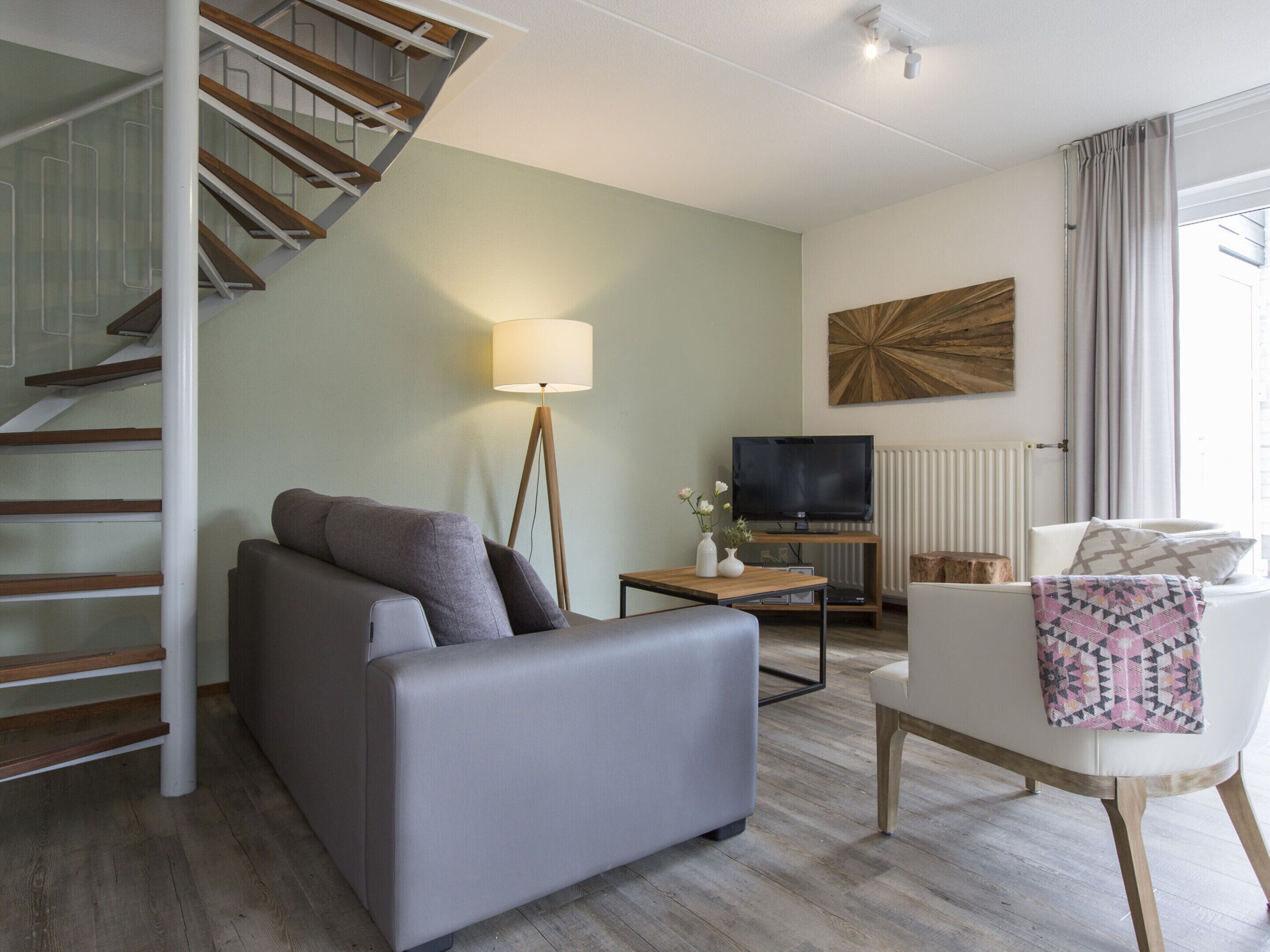 A modern living room with a grey sofa, a white armchair, a wooden coffee table, a floor lamp, a flat-screen TV, and a staircase with wooden steps.