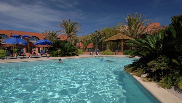 A swimming pool with clear water, surrounded by palm trees and umbrellas, with a building in the background. - Apartment in France Near Serene Beaches (Agde)